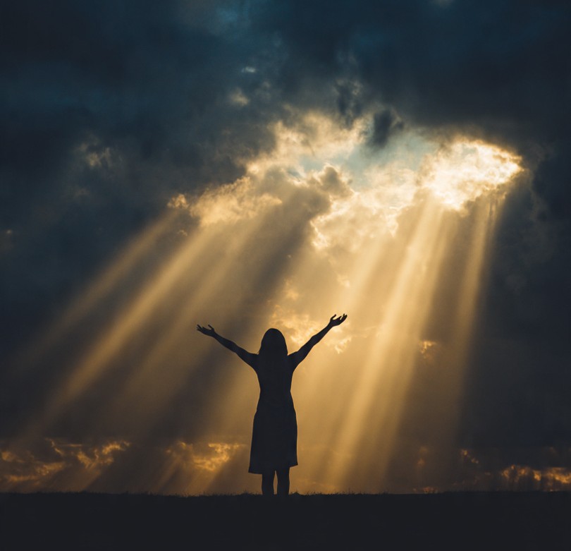 Woman with arms raised under light breaking through clouds